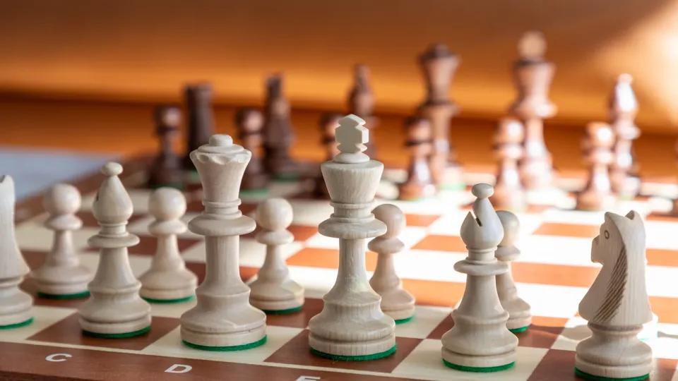 Primer plano de un tablero de ajedrez con piezas de madera: el rey blanco y su fila delante, y las negras desenfocadas al fondo, en tonos cálidos. Metáfora de anticipar jugadas.