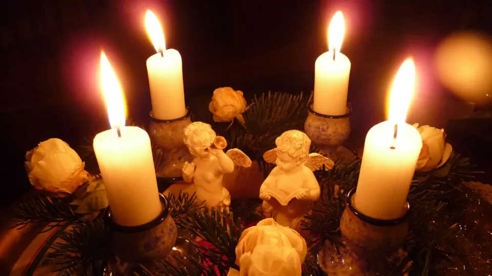 Corona de Adviento con cuatro velas encendidas, ramas de abeto, rosas secas y dos figuras de ángeles, en una sala a oscuras. Ambiente navideño; no es el estudio de Múnich.