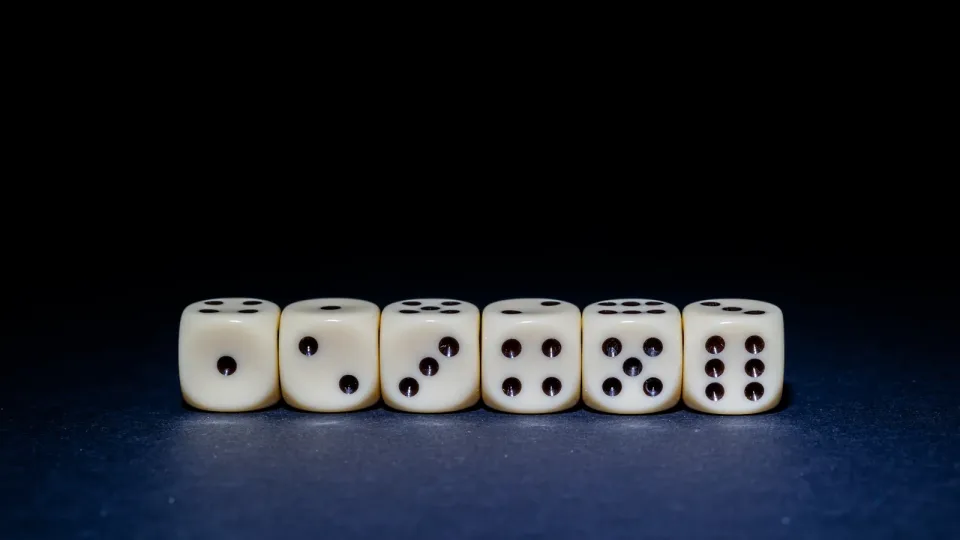 Seis dados de marfil en fila sobre una superficie azul oscura con fondo negro, mostrando las caras del uno al seis. Imagen de azar y probabilidad.